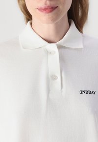 Biely polo tričko s textúrovanou tkaninou, dva gombíky na golieri a čiernym vyšitým logom "2NDDAY" na prednej strane.