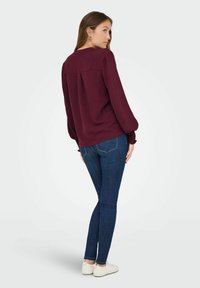 Karmīza garām piedurknēm ar teksturētu audumu, brīvā piegriezuma un kroku detaļām uz piedurknēm, apvienota ar tumši ziliem skinny džinsiem un baltām kurpēm.