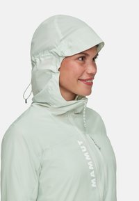 Giacca leggera verde menta con cappuccio aderente, cordini regolabili e zip frontale. Presenta un logo ben visibile sul petto.