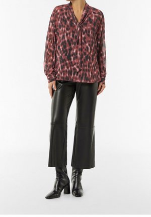 Kvinde iført en rød og sort mønstret bluse med bindebånd i halsen, sorte vidde læderbukser og sorte ankelstøvler med hæl.