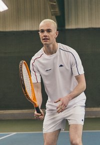 Valge tennisepaita, millel on punased ja sinised detailid, koos valgete lühikeste pükstega. Mängija hoiab oranži tennisereketit, valmis lööki sooritama.