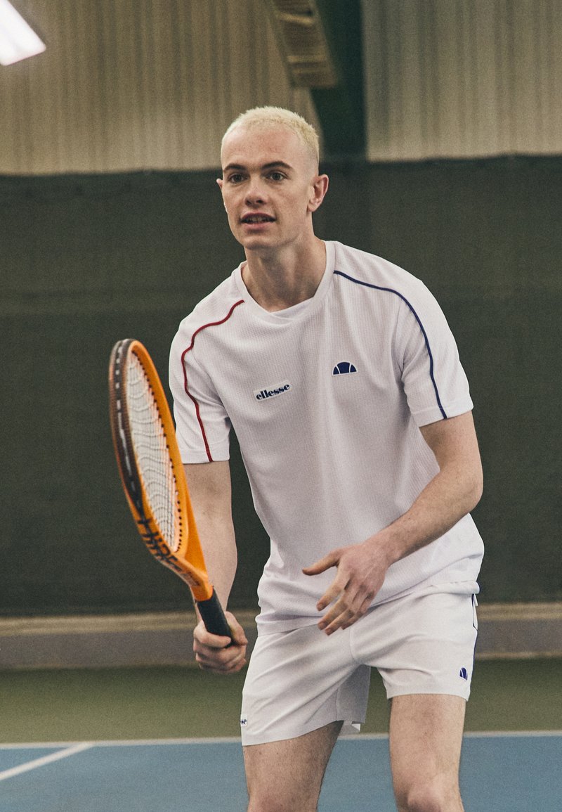 Valge tennisepaita, millel on punased ja sinised detailid, koos valgete lühikeste pükstega. Mängija hoiab oranži tennisereketit, valmis lööki sooritama.