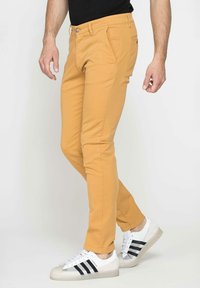 Pantaloni slim fit da uomo in giallo senape, realizzati in misto cotone. Presentano tasche anteriori e una chiusura con bottone classico. Abbinati a sneakers bianche.