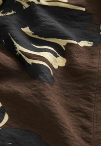 Primo piano di tessuto marrone con stampa floreale astratta in nero e beige e linea di cucitura visibile.