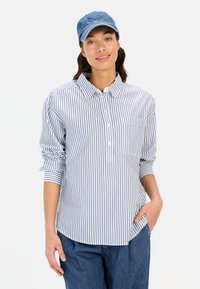Gestreifte Hemdbluse in Blau und Weiß, aus Baumwolle, mit Brusttasche und hochgerollten Ärmeln; kombiniert mit einer blauen Denim-Mütze.