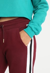 Teal kort sweatshirt med ribbestrikkede mansjetter, parret med burgunder joggebukser som har hvite og svarte sidestiper og snorstramming.