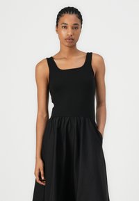 Zwarte ribgebreide tanktop met vierkante halslijn, gecombineerd met een zwarte vloeiende rok met een geplooide taille. De outfit heeft een soepele textuur.