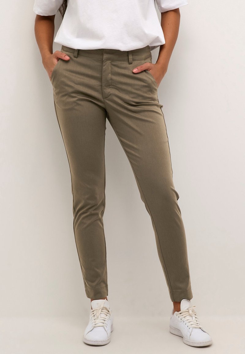 Femme portant un pantalon slim vert olive et des baskets blanches, debout avec les mains dans les poches sur un fond uni.