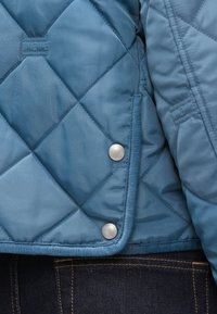 Close-up af blå quiltet jakke med sølvfarvede Tommy Hilfiger trykknapper og synlige sting, båret over mørke denim jeans.