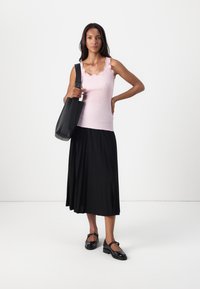 Débardeur rose à côtes avec des bords festonnés, associé à une jupe plissée noire. Le mannequin porte un sac à main noir et des chaussures Mary Jane noires.