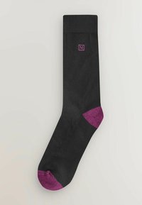 Musta sukka, jossa on teksturoitu pinta ja vaaleanpunainen yksityiskohta varpaassa ja kantapäässä. Yläosassa pieni violetti logo.