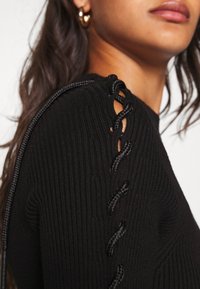 Haut en maille côtelée noire avec un détail à lacets sur l'épaule, en tissu texturé et à la coupe ajustée.