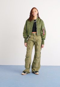 Olijfgroene bomberjack met een cropped ontwerp, gecombineerd met olijfgroene cargo broeken met meerdere zakken. Zwarte t-shirt eronder, zwart en tan sneakers.