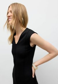 Robe noire ajustée avec un décolleté en V et des manches courtes, ornée de coutures verticales. Un bracelet de couleur claire orne le poignet. Fond uni.