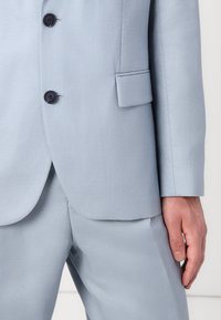 Traje ajustado de color azul claro con una textura suave, que presenta dos botones oscuros, un bolsillo con solapa y pantalones bien planchados.