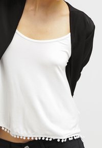 Cardigan nero sopra una canotta bianca. La canotta presenta un orlo ondulato con piccole decorazioni a pompon. Tessuto con una finitura liscia.