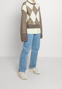 Brun og creme argyle sweater med lange ærmer, lagd ovenpå en lys grøn skjorte, parret med blå jeans og hvide sneakers.
