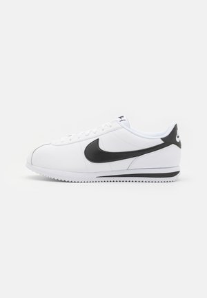 White leather sneakers with black Nike swoosh, flat laces, and textured rubber sole. Rounded toe and padded collar for comfort.