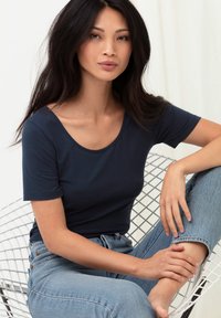 Námořnická modrá blůza s krátkým rukávem a kulatým výstřihem, zkombinovaná se světle modrými džíny. Model sedí na bílé drátěné židli.