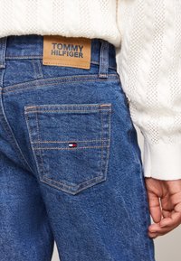 Vaqueros de mezclilla con un lavado azul oscuro, que cuentan con una etiqueta de cuero con la inscripción "TOMMY HILFIGER" en la cinturilla y un pequeño logo tricolor en el bolsillo trasero.