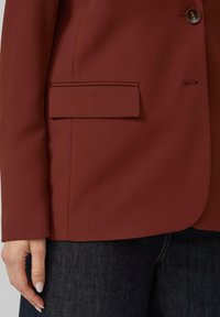Linkerzijde van een roestkleurige blazer met een klepzak, een bruine knop en de hand van een persoon die rust dichtbij donkere denim jeans.