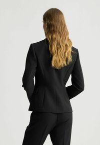Blazer nero su misura con spalle strutturate, che mostra una silhouette elegante. Presenta un tessuto testurizzato e tasche laterali doppie.