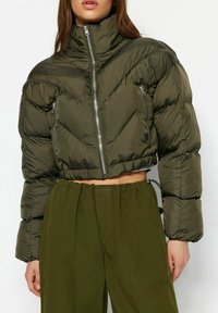 Žena oblečená v oříznuté péřové bundě olivově zelené barvy zapnuté na zip, doplněná o kalhoty s vysokým pasem stejné barvy, na jednobarevném pozadí.