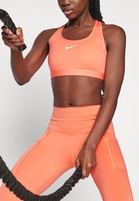Nike sportski grudnjak i tajice visokog struka u vibrantnoj koralj boji, izrađene od rastezljivog materijala, s bijelim logotipom i džepovima na tajicama.