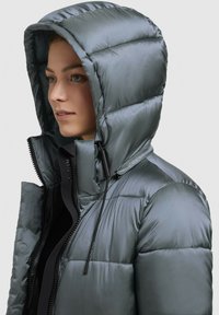 Chaqueta acolchada en tejido gris metálico con capucha, cierre de cremallera y cordón ajustable. Presenta un diseño acolchado y una textura suave.