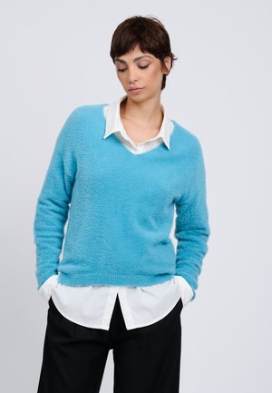 Mujer con cabello castaño corto mirando hacia abajo, que lleva un suéter azul claro y esponjoso sobre una camisa blanca con cuello, con pantalones negros y las manos en los bolsillos.