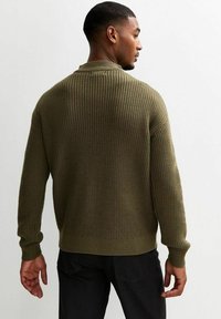 Olivgrön stickad tröja med ribbat mönster, långa ärmar och rund halsringning, tillsammans med svarta byxor.