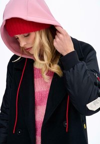 Žena si upravuje růžovou kapuci přes červenou čepici, má na sobě černou bundu s červenými šňůrkami a růžový pruhovaný pletený svetr na bílém pozadí.