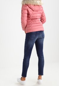 Vadderad rosa jacka med huva i fuskpäls, ribbad struktur och figursydd design, kombinerad med mörkblå jeans och vita sneakers.