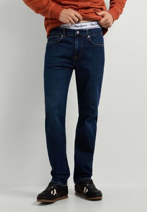 Person trägt dunkelblaue Jeans, schwarze Sneakers und ein orangefarbenes Langarmshirt und richtet den Bund so, dass das weiße Unterwäscheband von Pepe Jeans sichtbar wird.