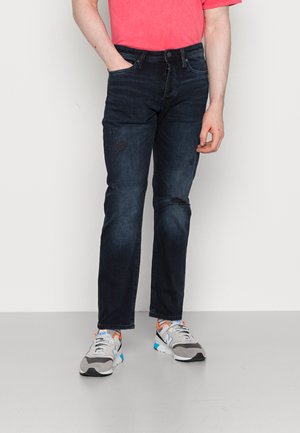 Σκούρες μπλε τζιν με χαλαρή εφαρμογή, με φθαρμένες λεπτομέρειες στο γόνατο και συνδυασμένες με γκρι και πορτοκαλί sneakers.