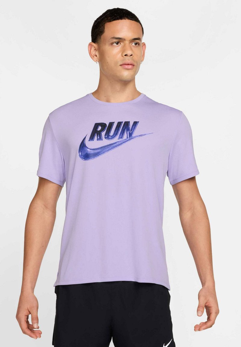 Homme portant un t-shirt Nike lavande avec le mot « RUN » et le logo swoosh en bleu, associé à un short Nike noir, faisant face à l'avant avec une expression neutre.
