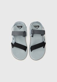 Sandales grises avec deux brides noires réglables, une semelle texturée et un logo sur la bride supérieure. Design plat adapté à un usage décontracté.