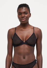 Svart bralette med gjennomsiktig prikket stoff, trekantede kopper, tynne stropper og bølget kant langs kantene. Justerbar baklukking.