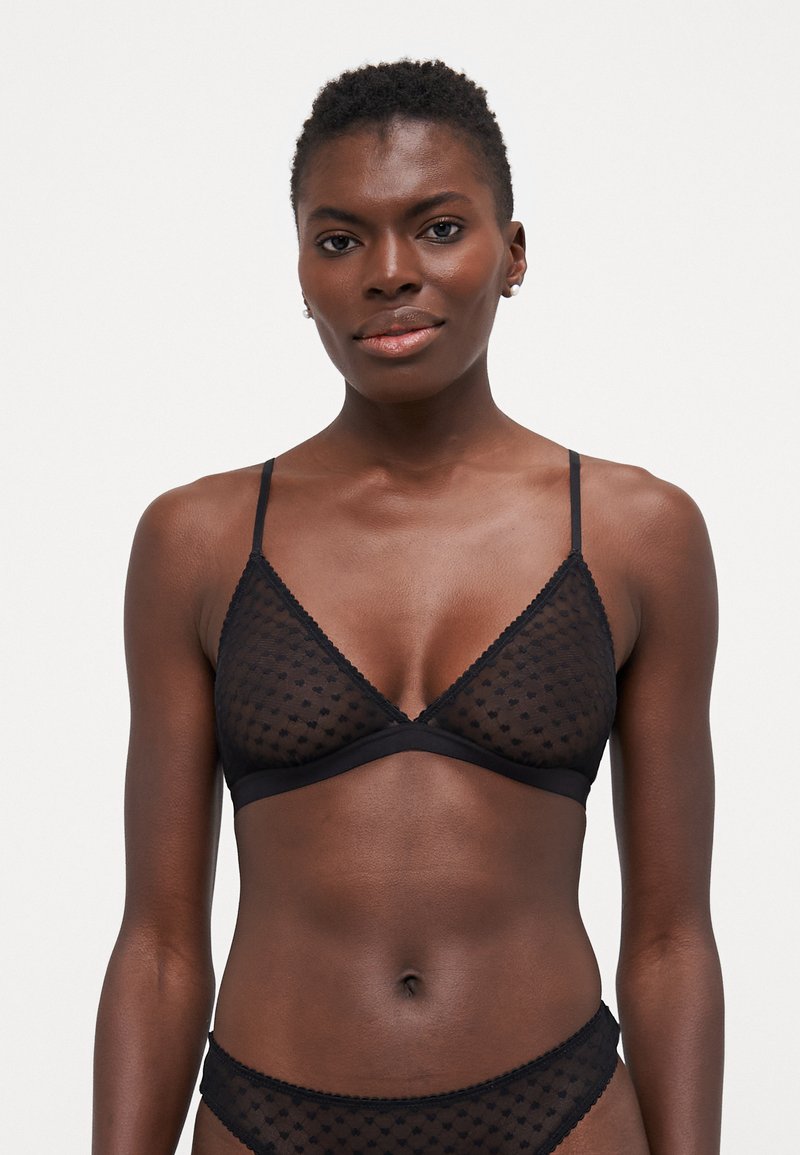 Svart bralette med gjennomsiktig prikket stoff, trekantede kopper, tynne stropper og bølget kant langs kantene. Justerbar baklukking.