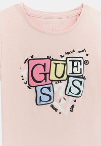 Rožinė organinės medvilnės marškinėliai su daugiaspalve grafika, kurioje didžiosiomis raidėmis parašyta „GUESS“, apsupta širdelių ir žaismingų teksto akcentų.