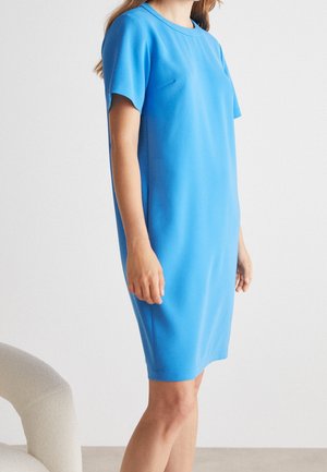 Femme portant une robe bleu vif, à manches courtes et longueur genou, sur un fond blanc uni, debout à côté d'une chaise beige incurvée.