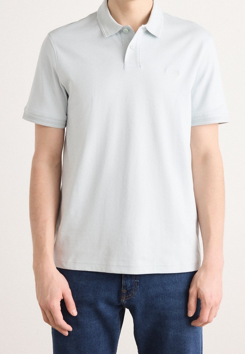 Homme portant un polo uni gris clair à manches courtes et un jean bleu foncé, debout devant un fond neutre.