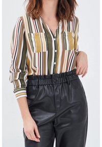 Chemise à boutons rayée en vert, jaune et blanc, avec deux poches poitrine et des manches longues. Portée avec un pantalon taille haute en cuir noir.