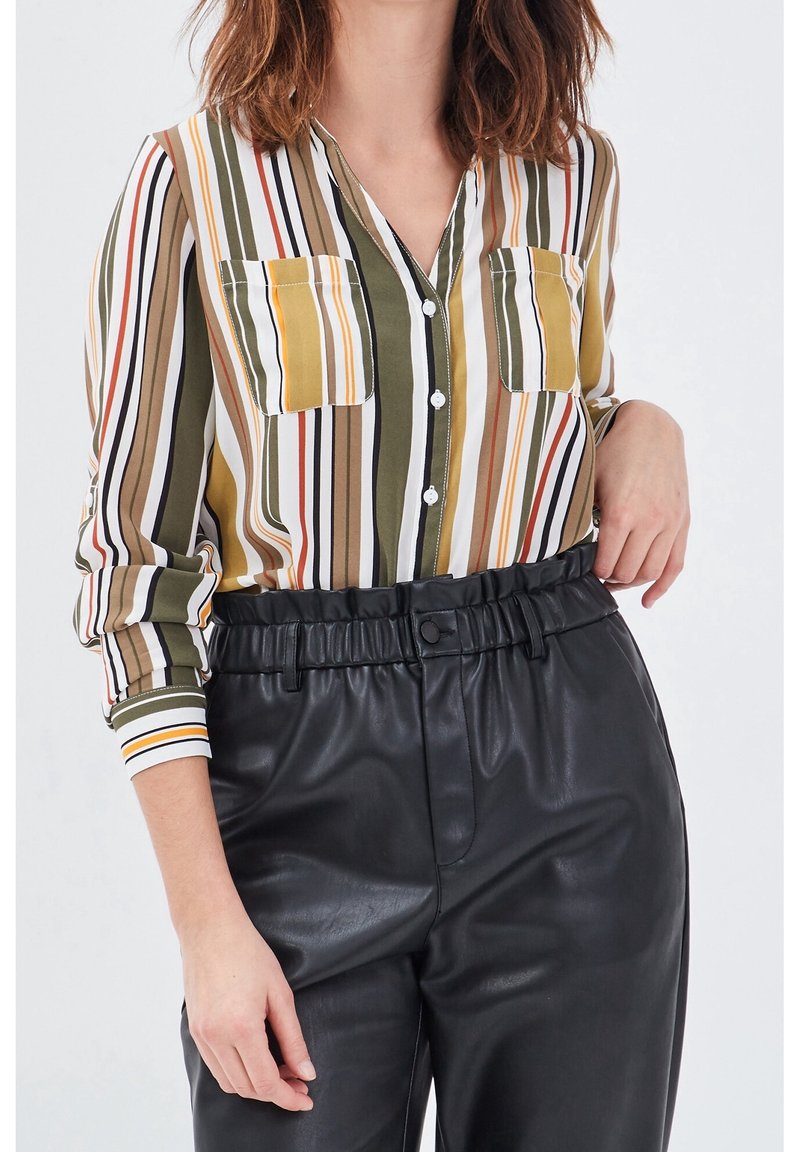 Chemise à boutons rayée en vert, jaune et blanc, avec deux poches poitrine et des manches longues. Portée avec un pantalon taille haute en cuir noir.