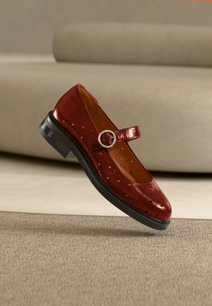 Piros bőr Mary Jane cipő perforált mintákkal és ezüst csattal, levegőben megjelenítve, texturált bézs felület felett.