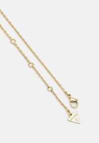 Collar de cadena en tono dorado con un colgante triangular que presenta el logo de "Guess", cierre de langosta y enlaces decorativos. Textura de metal suave.