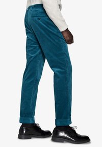 Teal corduroy-housut suoran leikkauksen kanssa, joissa on takatasku ja lahkeet. Yhdistettynä mustiin nahkakenkiin täydentämään asua.