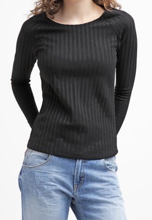 Femme portant un haut noir côtelé à manches longues ajusté et un jean bleu clair délavé, debout avec les mains derrière le dos.