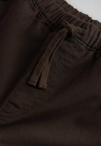 Tumši brūnas auduma bikses ar elastīgu vidukli un sasienamu auklu priekšējā kabatas apvidū.