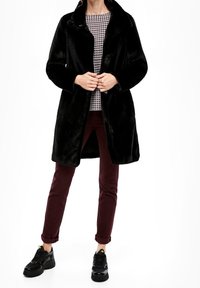 Sort kunstlæderfrakke med krave, fortil knapper, båret over en mønstret top. Sammen med burgundy bukser og sorte sneakers.